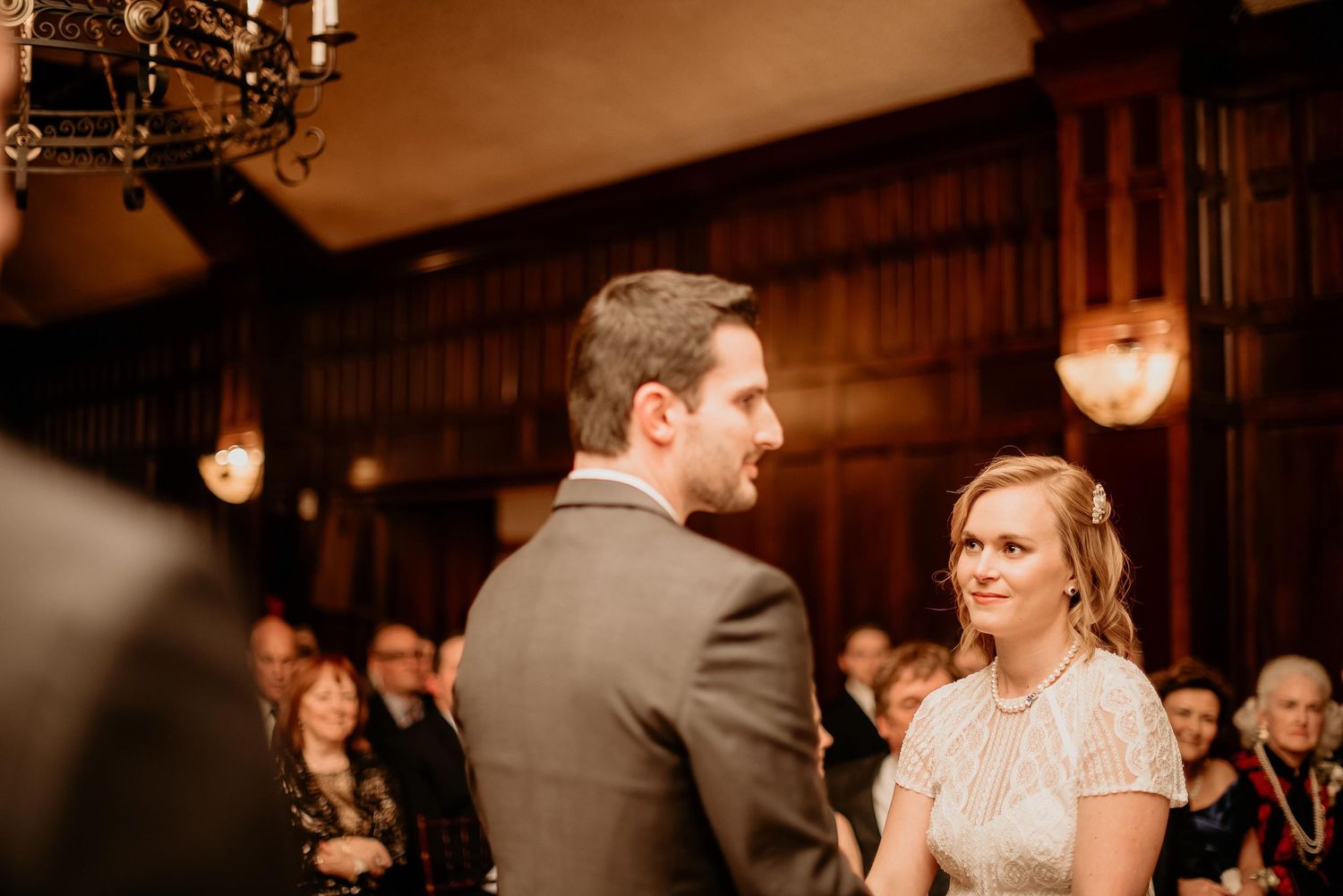 Bride and groom exchange vows at elegant wood-paneled wedding ceremony with guests seated in background.