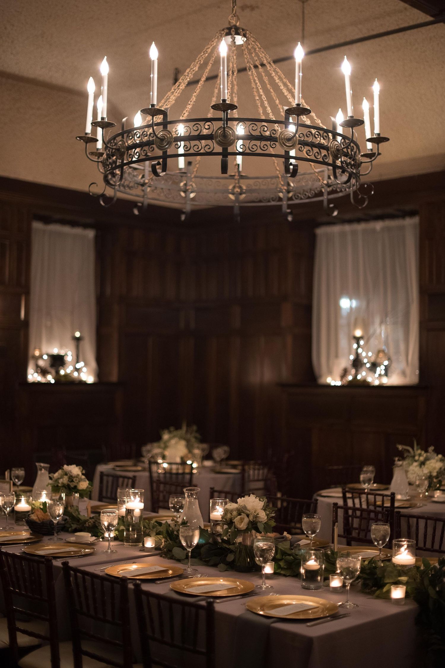 Elegant wedding reception with ornate iron chandelier, candlelit tables, gold chargers, floral centerpieces, and dark wood paneled walls.