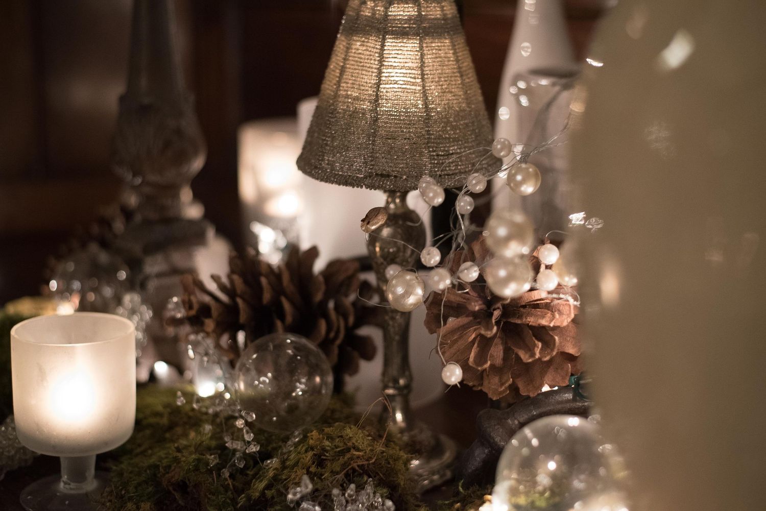 Elegant Christmas table display with glowing candles, pine cones, fairy lights, and a small decorative lamp.