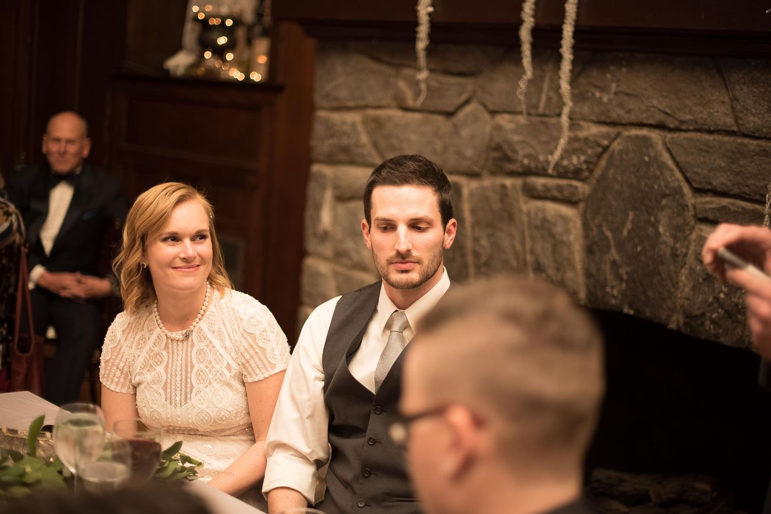 Bride and groom seated together at wedding reception near stone fireplace in elegant rustic venue.
