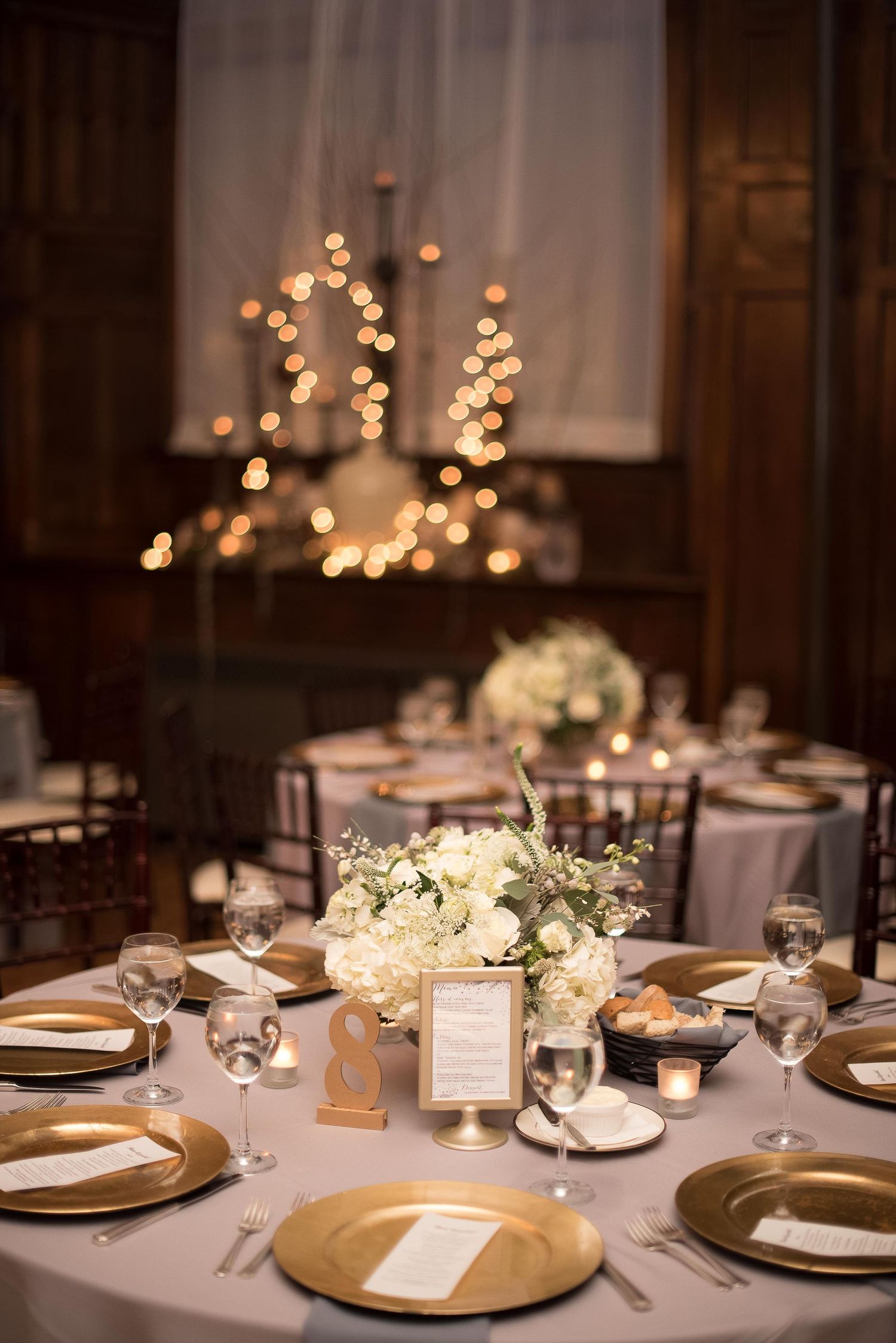 Elegant wedding reception table with gold charger plates, white floral centerpiece, and soft candlelight in a warmly lit venue.