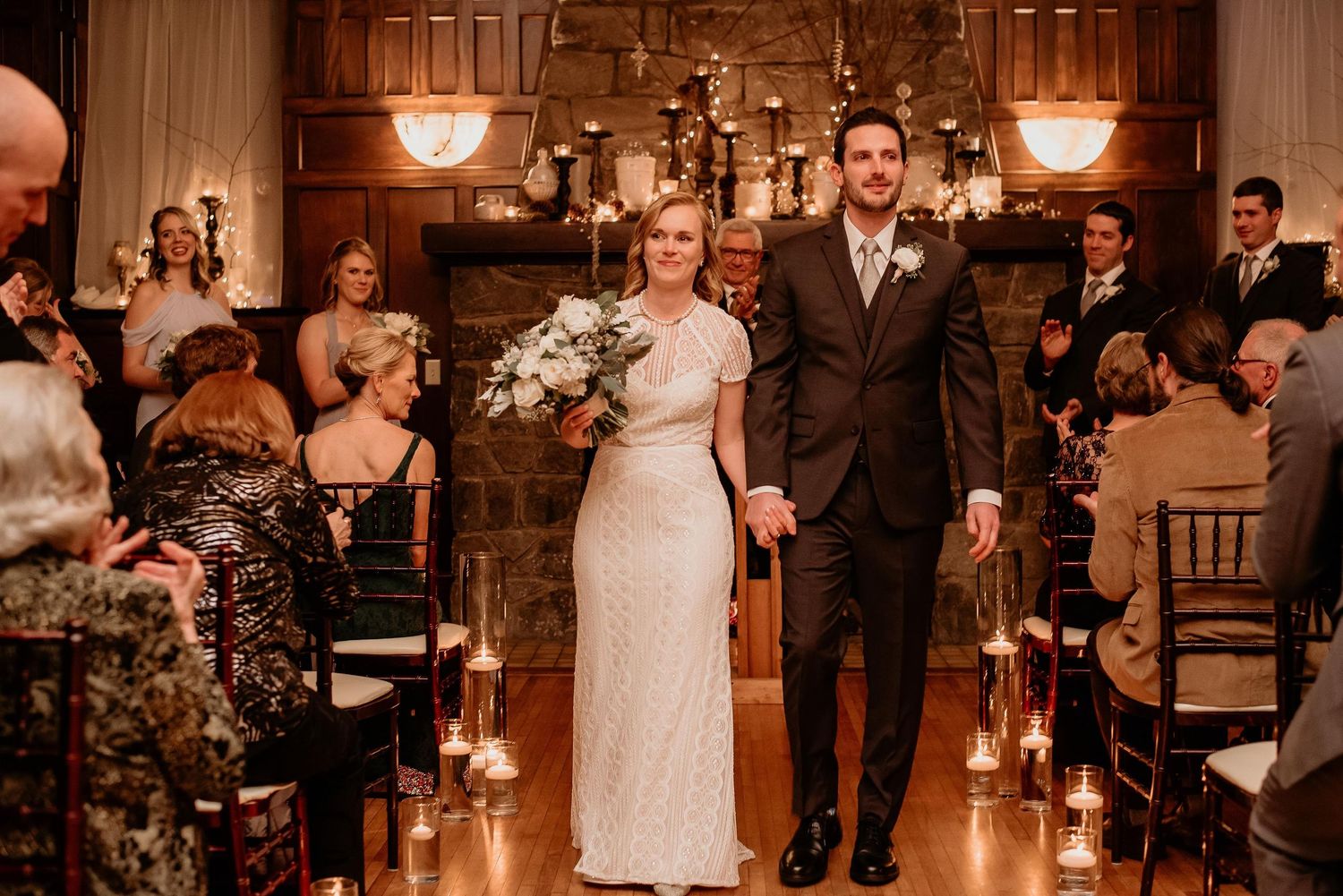 Bride and groom walk down aisle at candlelit indoor wedding ceremony with stone fireplace backdrop.