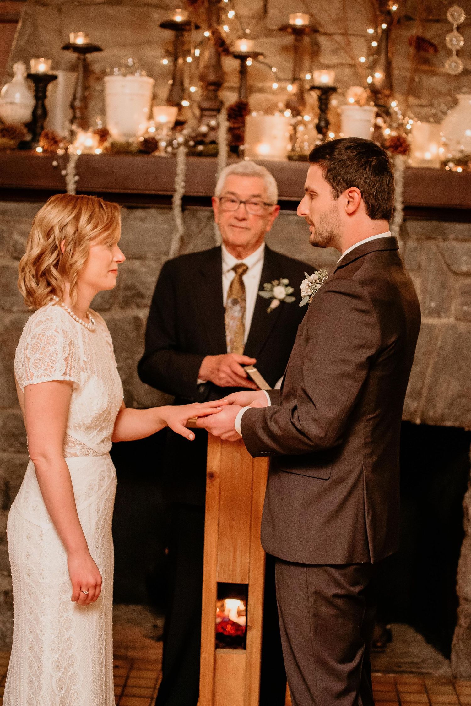 Bride and groom exchange rings at cozy indoor wedding ceremony by candlelit stone fireplace with officiant.