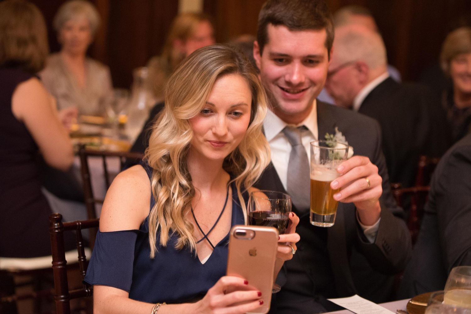Couple smiling at formal dinner event, woman taking selfie with iPhone, man holding beer glass.