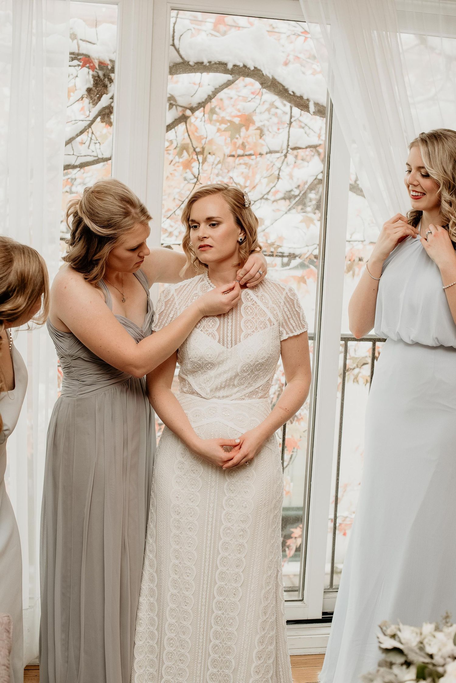 Bridesmaids helping bride get ready in white lace gown near floral window backdrop.