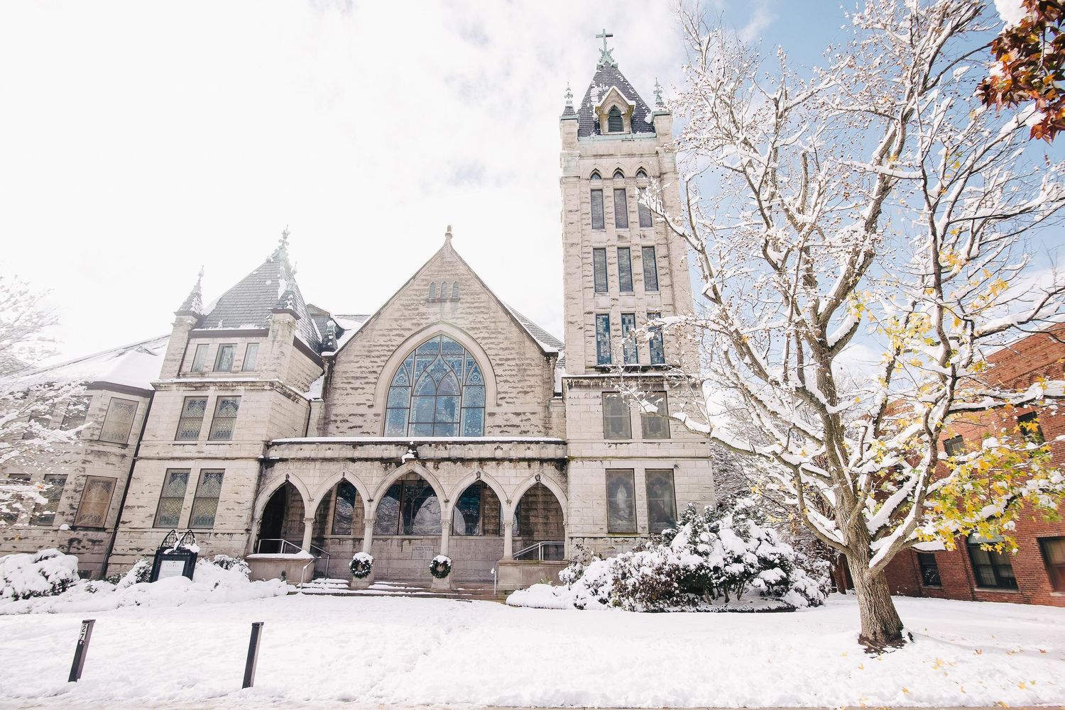 Snow-covered Gothic stone church with bell tower and bare winter trees under overcast sky.