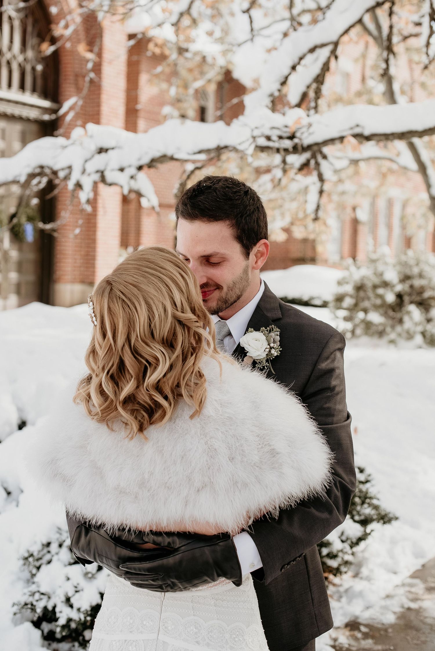 Bride and groom share a romantic winter wedding moment outdoors, surrounded by snow-covered trees and a brick building.