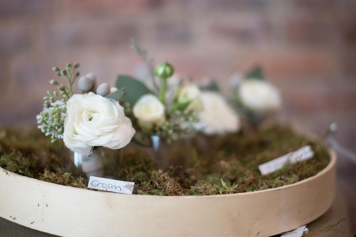 White ranunculus boutonnieres displayed on a moss-covered wooden tray with eucalyptus greenery for a rustic wedding.