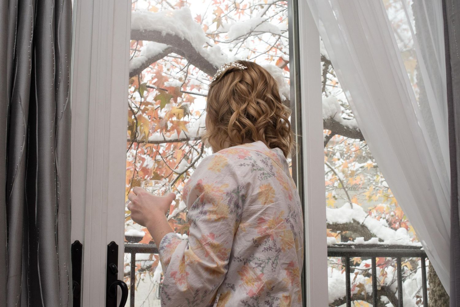 Woman in floral robe gazing out open window at autumn trees with snow-dusted branches.