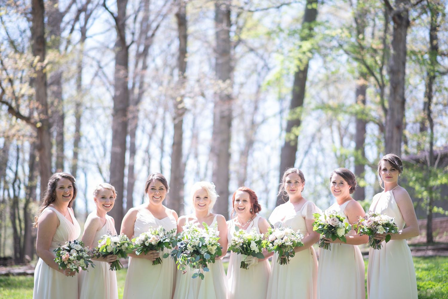 Eight bridesmaids in ivory gowns holding white and green bouquets in wooded setting.