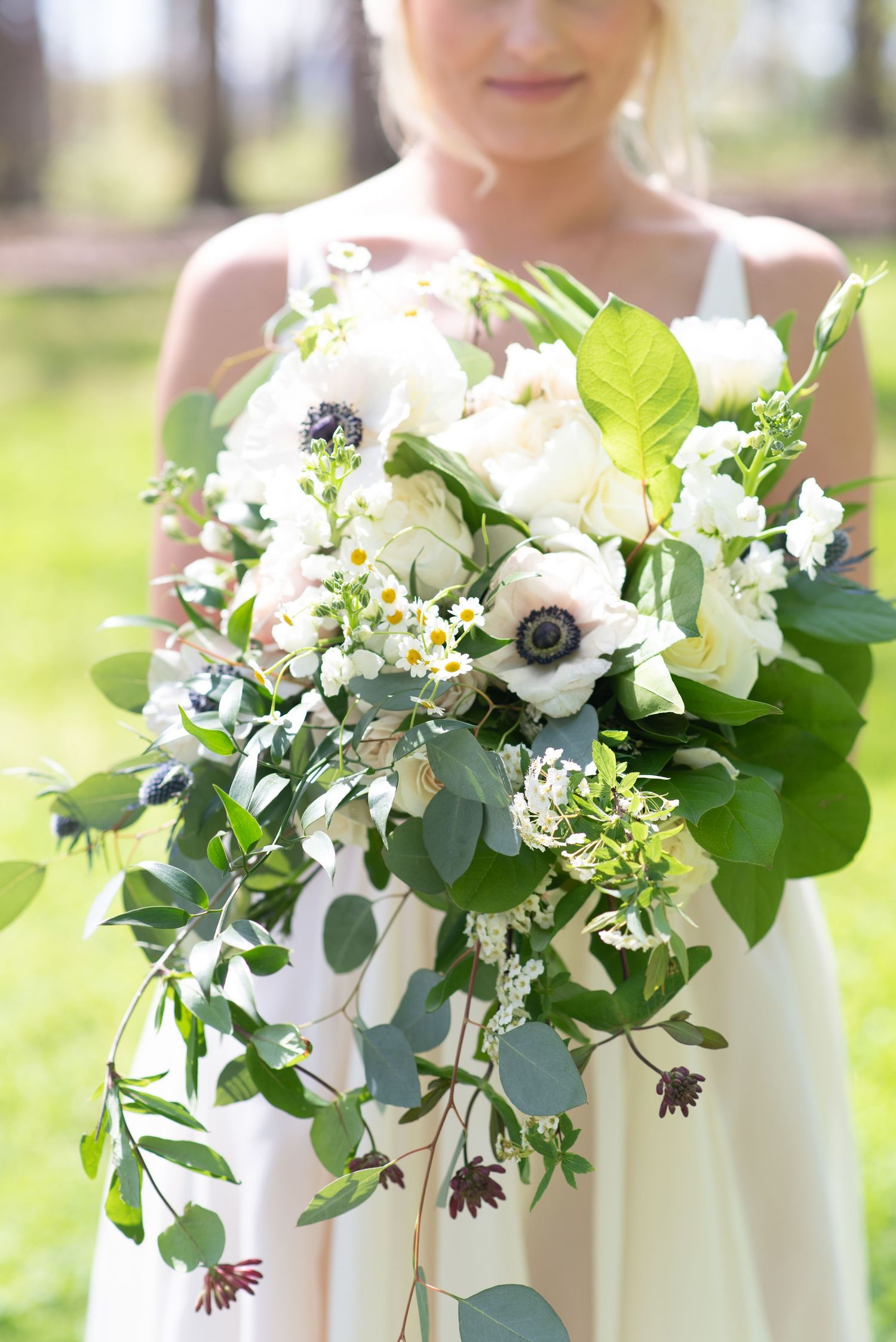Lush cascading bridal bouquet with white anemones, roses, and greenery held by bride in white dress.