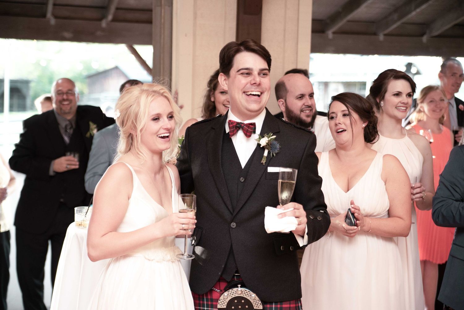 Bride and groom in Scottish kilt celebrate with champagne alongside bridesmaid at wedding reception.