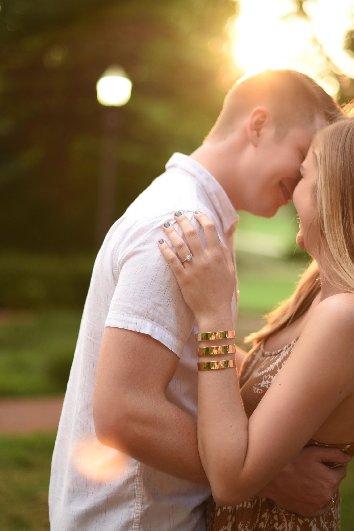 Young couple touching foreheads tenderly at golden sunset in park, woman wearing gold bracelets.