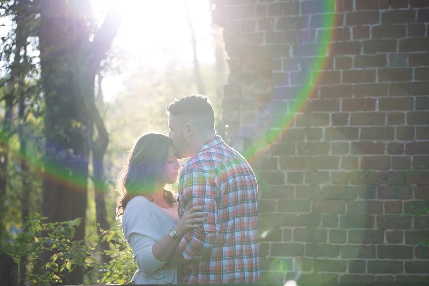 A couple shares a romantic moment near a brick wall with beautiful sun flare filtering through the trees.