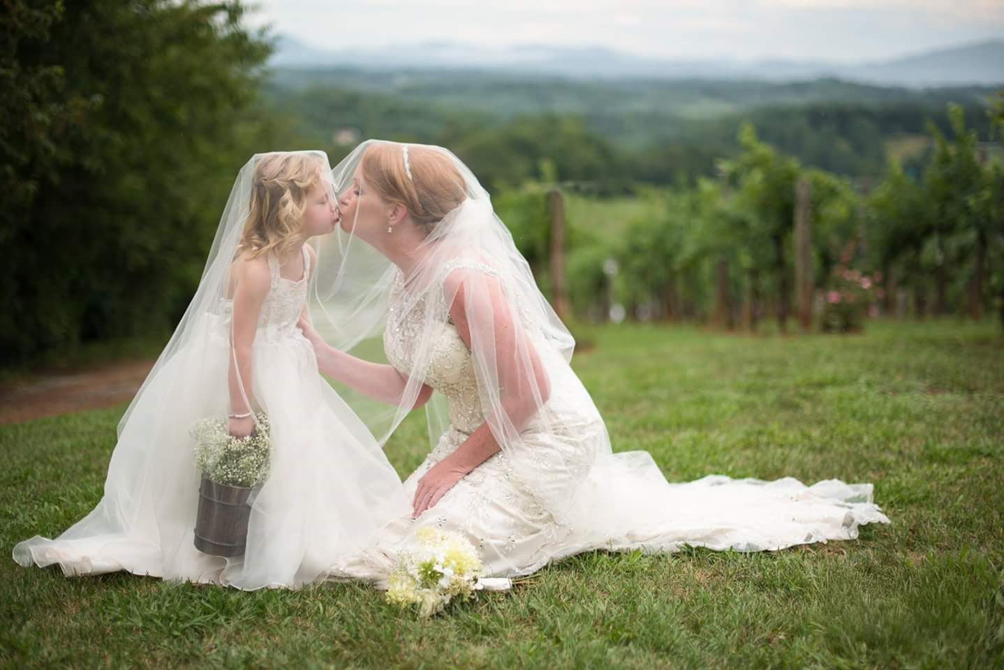 Two brides in white gowns and veils share a kiss while sitting on green grass at a vineyard wedding.