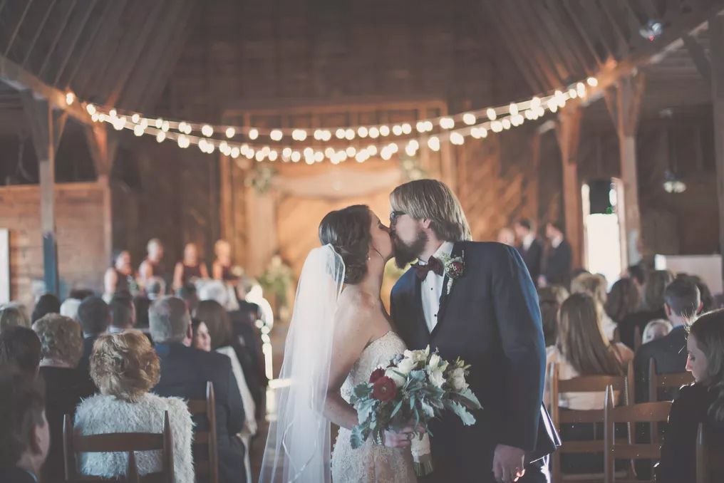 Bride and groom share first kiss at rustic barn wedding ceremony with string lights and seated guests.
