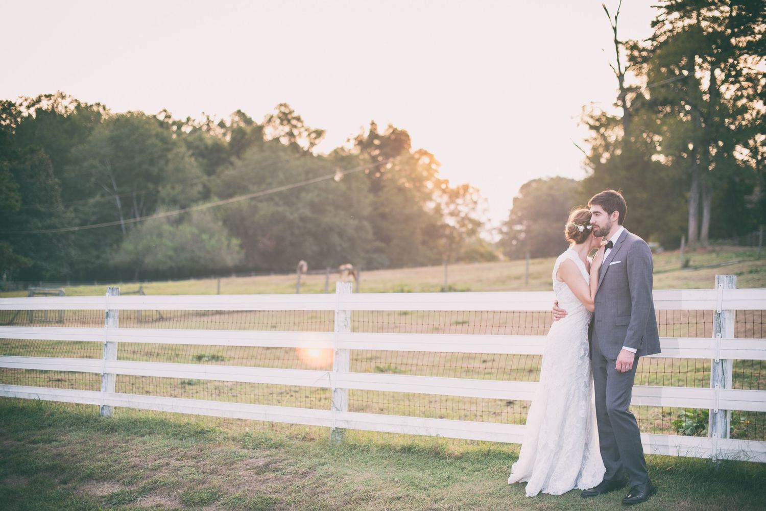 Bride and groom share a tender moment by a white fence at golden hour sunset.