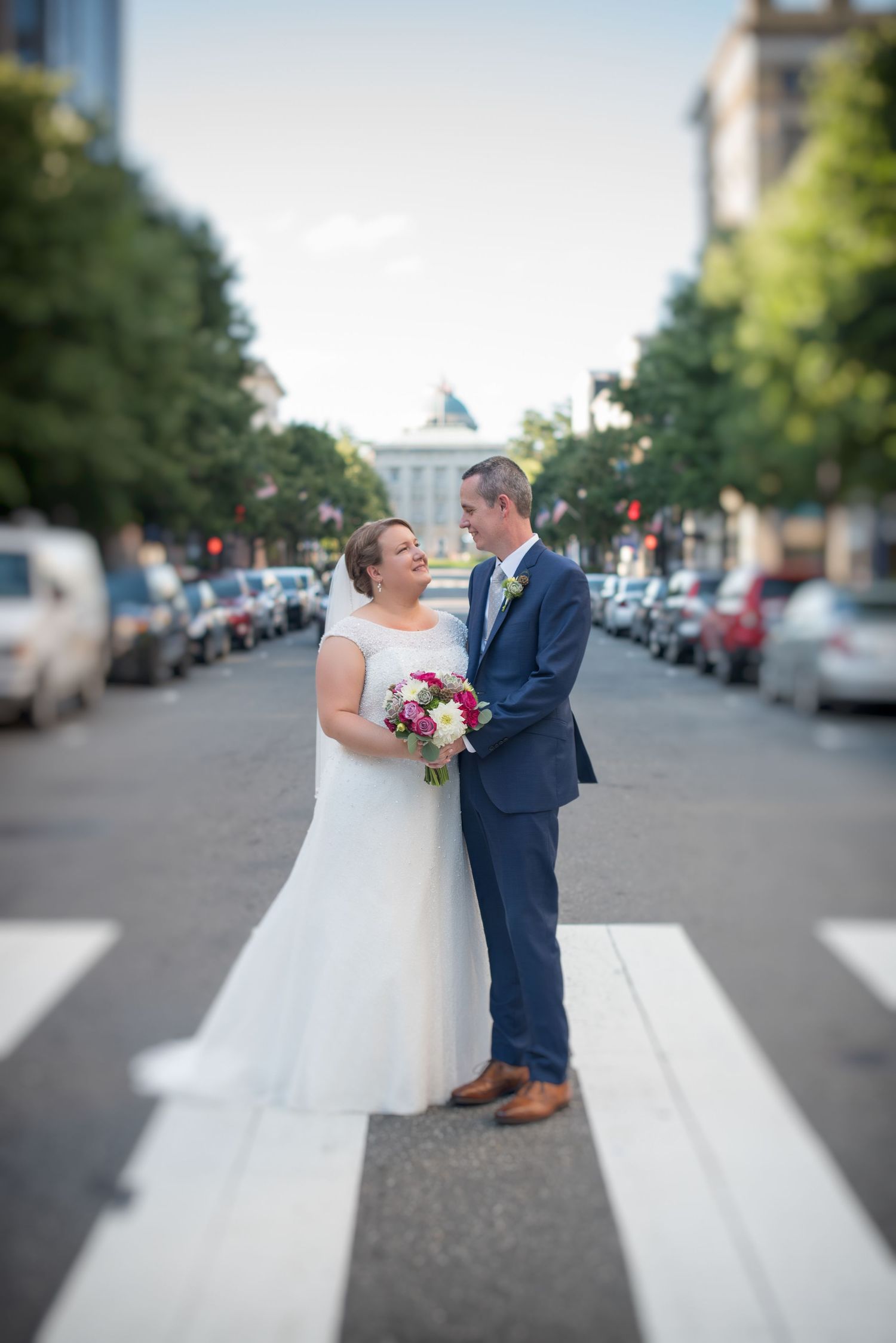 Bride and groom share a romantic moment on a city crosswalk, bride holding a pink and white bouquet.
