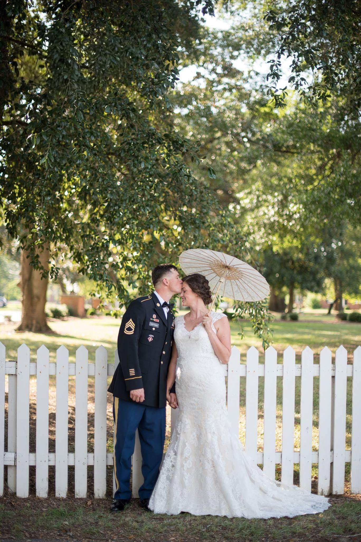Military groom in dress uniform kisses bride holding parasol beside a white picket fence outdoors.