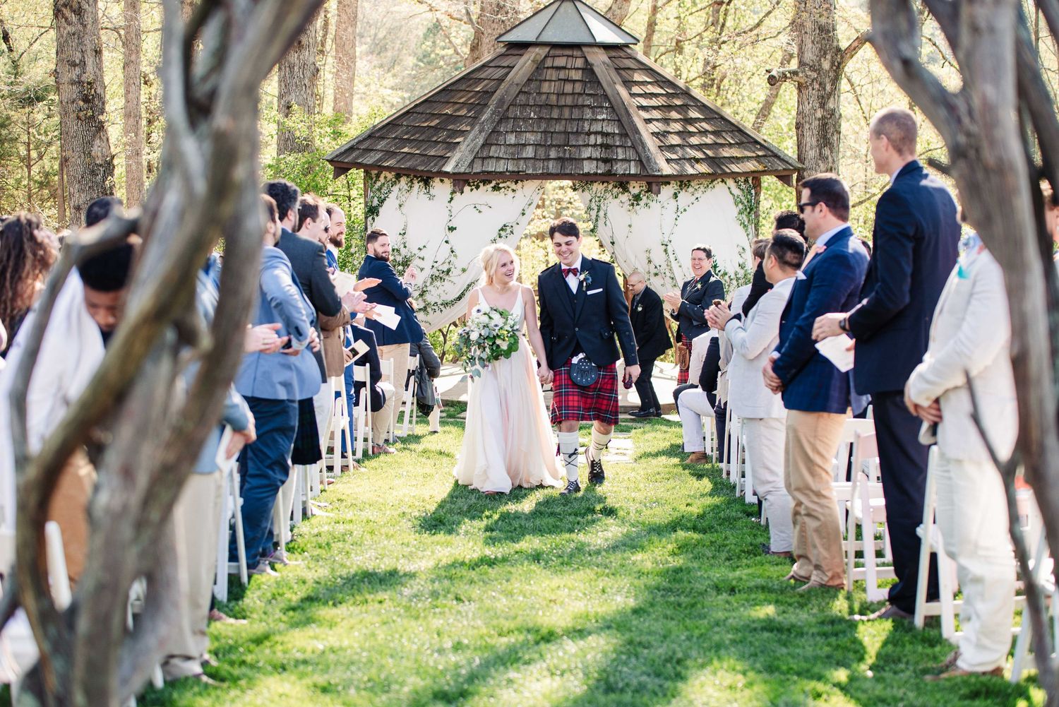 Outdoor wedding ceremony with bride and groom walking down aisle past guests, gazebo backdrop, lush green trees, sunny day.