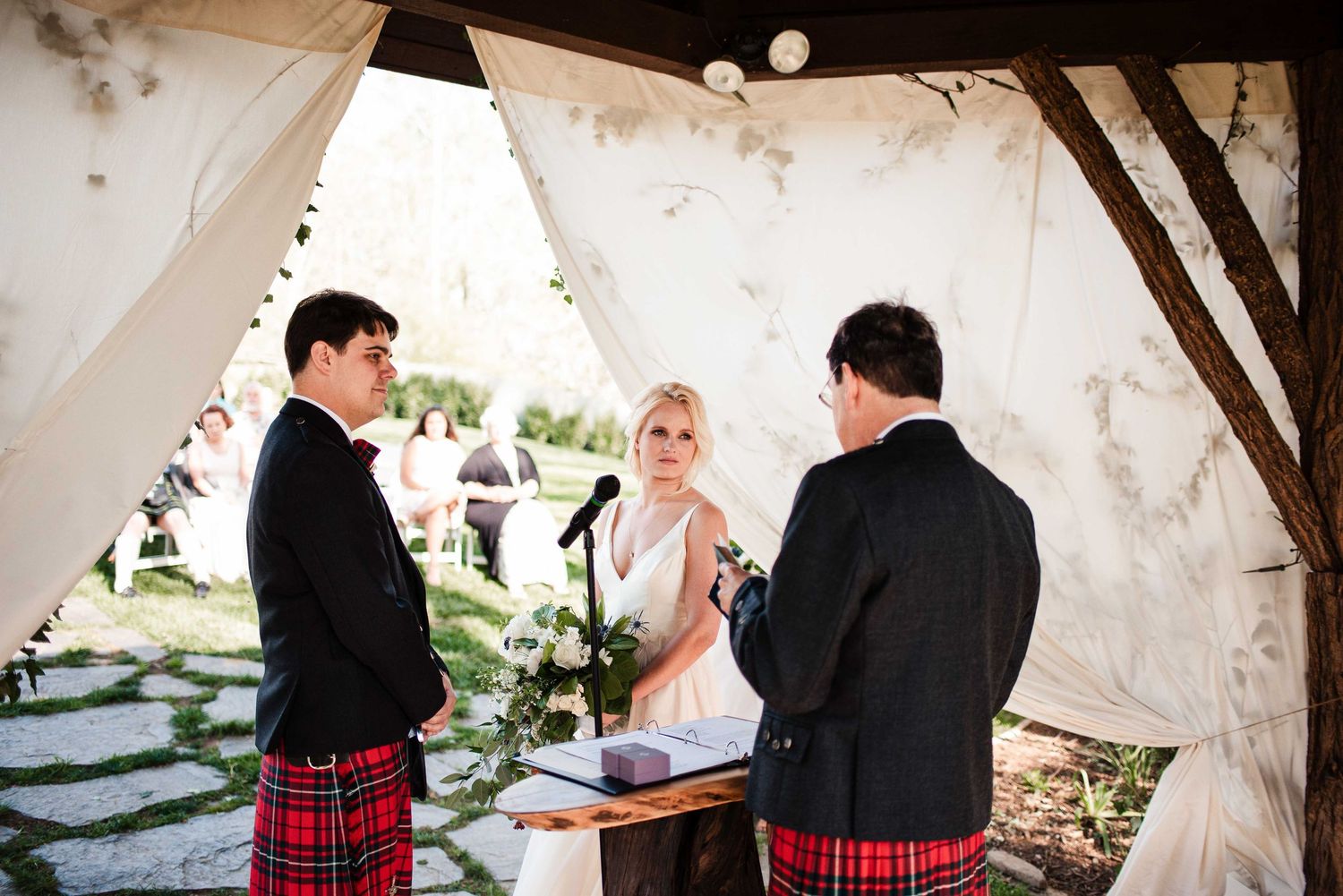 Bride and groom in red tartan kilt exchange vows with officiant under rustic outdoor wedding canopy.