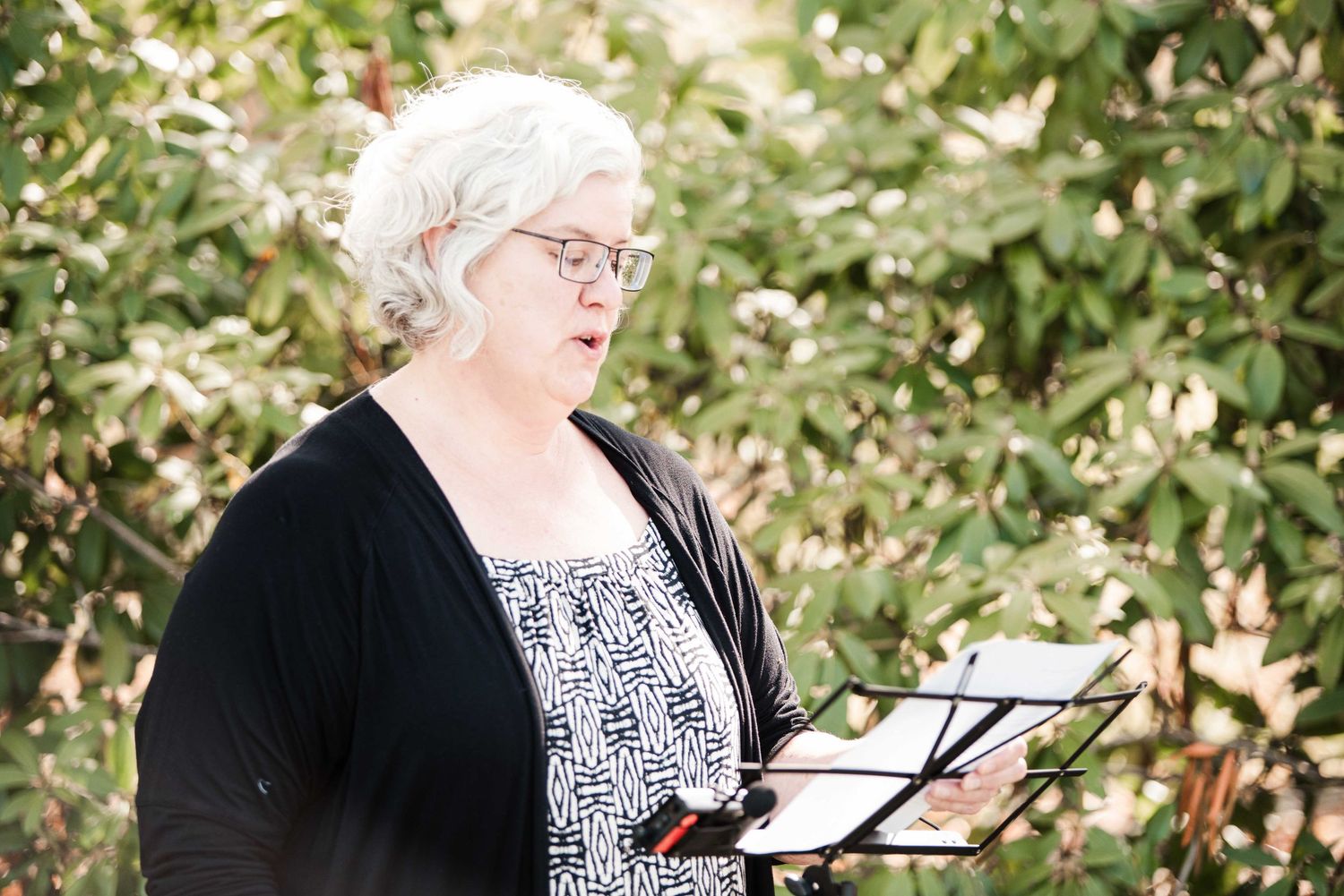 Woman with white hair and glasses reads from a music stand outdoors among green foliage.
