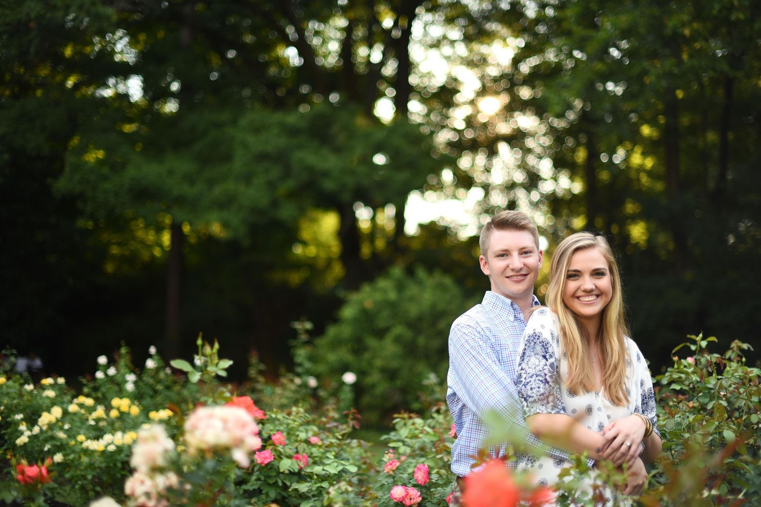 Couple smiling together in a lush garden surrounded by colorful roses and green trees in soft golden light.