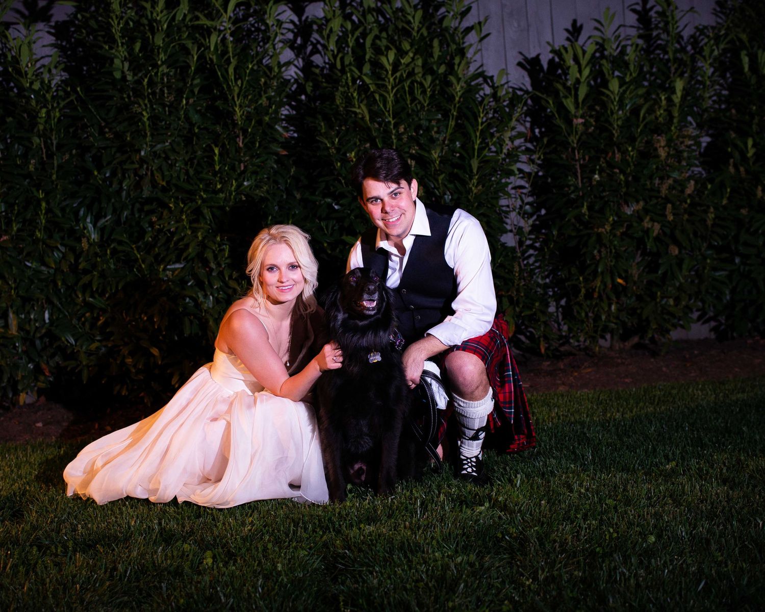 Bride and groom in Scottish kilt pose with black dog on grass at night wedding celebration.