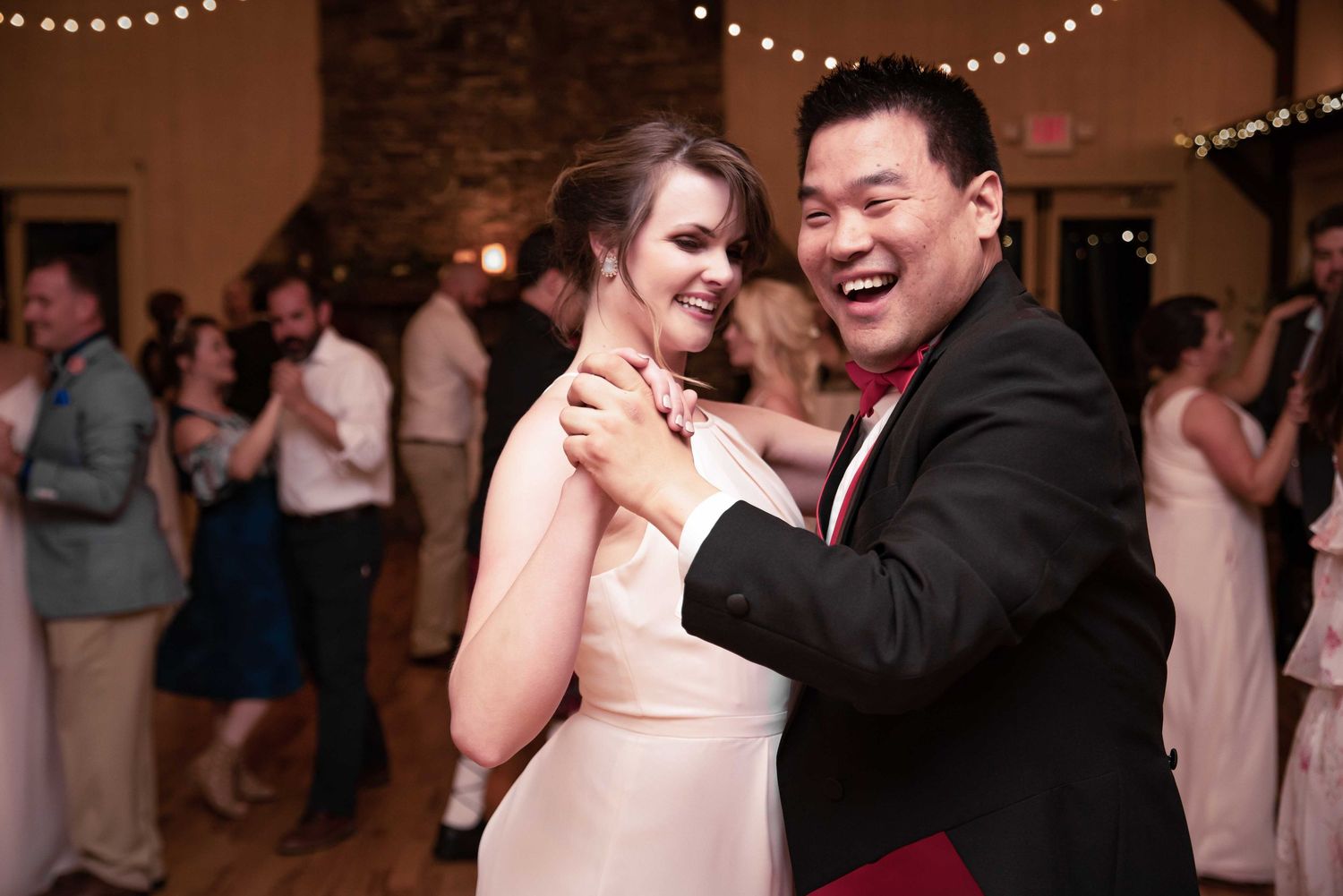 Couple dancing joyfully at a wedding reception in a rustic venue with string lights.