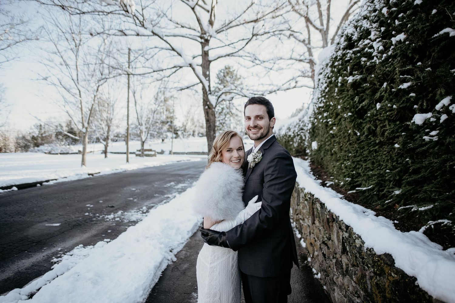 Happy couple hugging in snowy winter setting, bride in white gown with fur shawl, groom in dark suit outdoors.