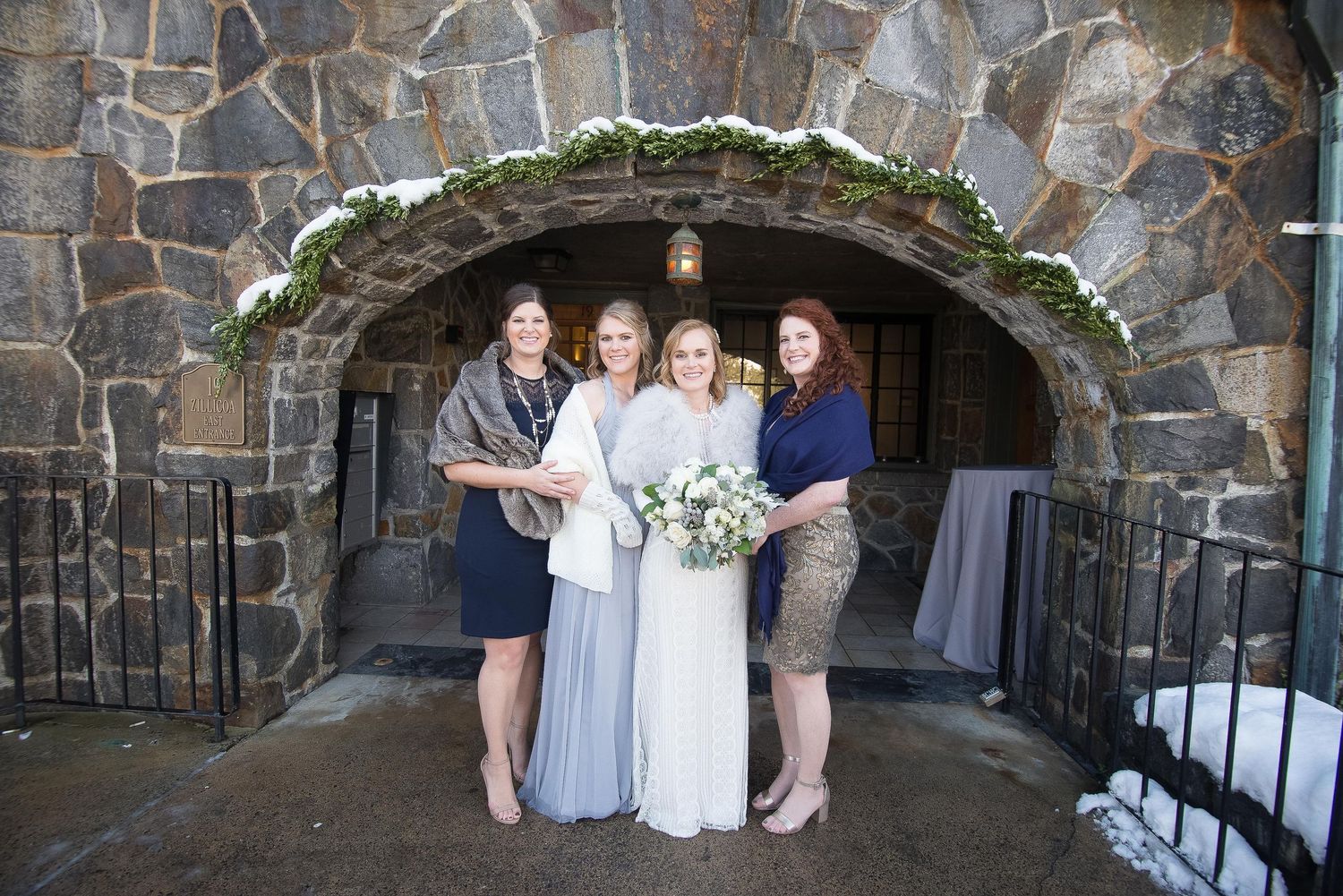 Bride with bouquet and three bridesmaids posing under stone arch decorated with greenery at winter wedding venue.