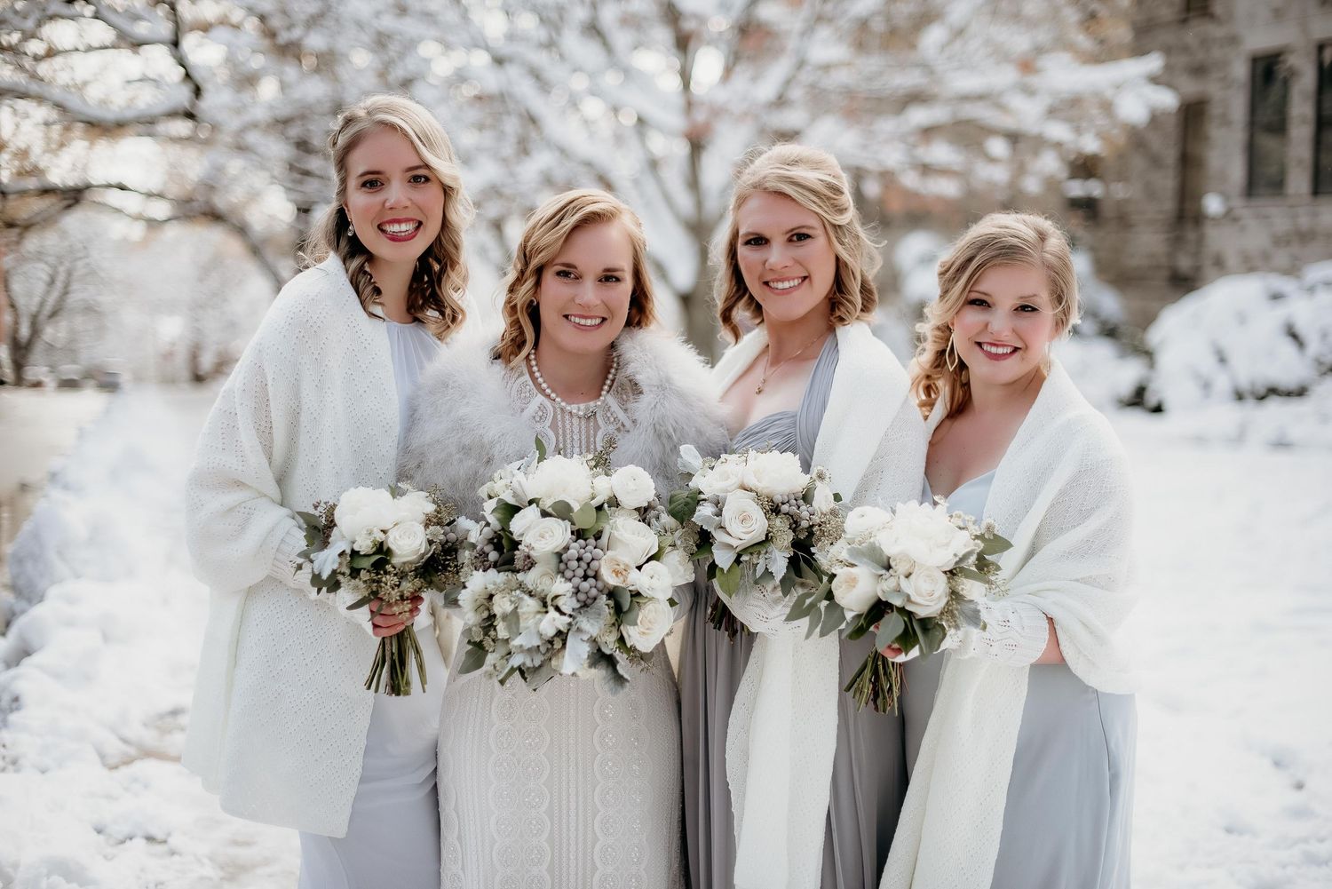 Four women in white wraps holding white bouquets pose together in a snowy winter wedding photo.