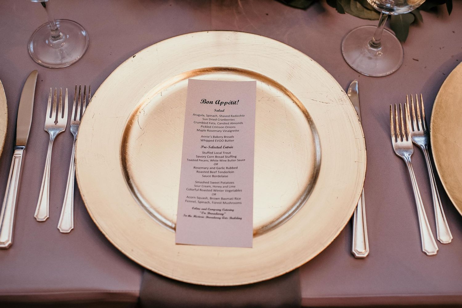Elegant wedding place setting with gold charger plate, silver cutlery, crystal glasses, and a printed menu card.