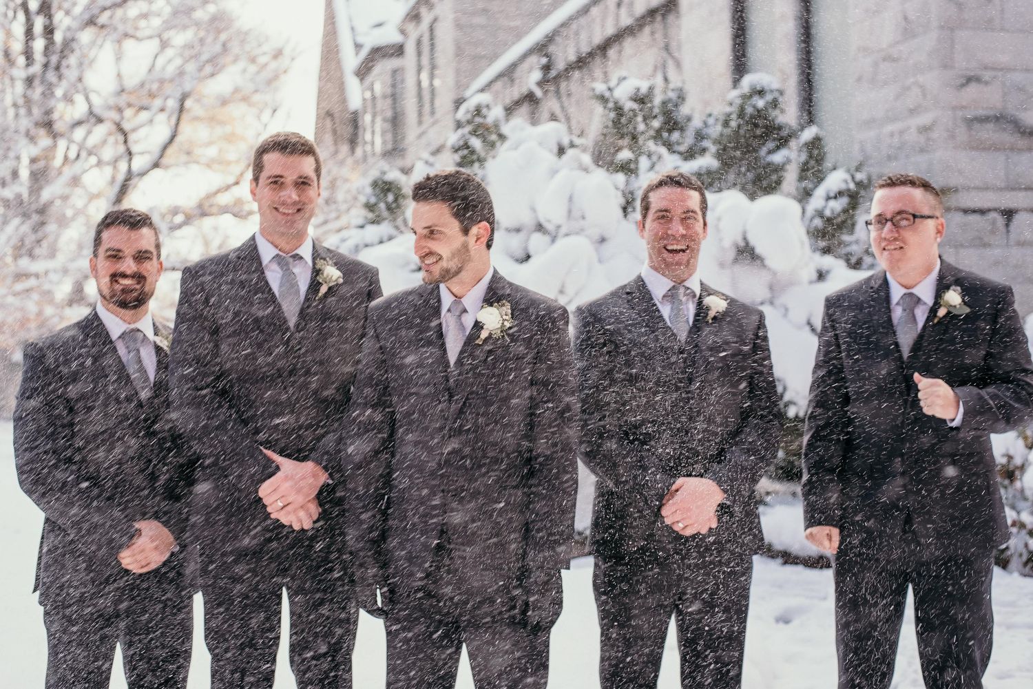 Five groomsmen in dark suits laughing together outdoors during a snowy winter wedding ceremony.