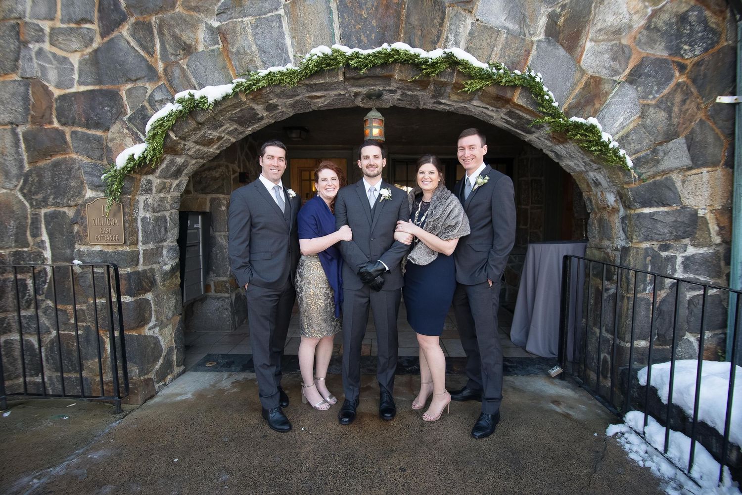 Five formally dressed guests posing together under a stone archway at a winter wedding venue.