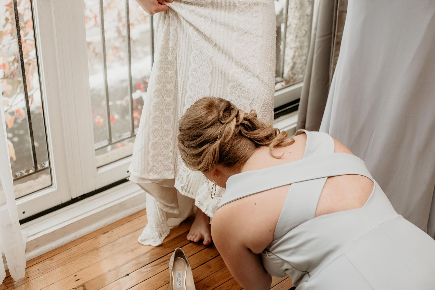 Bridesmaid kneeling to help bride into lace wedding dress near sunlit windows on hardwood floor.