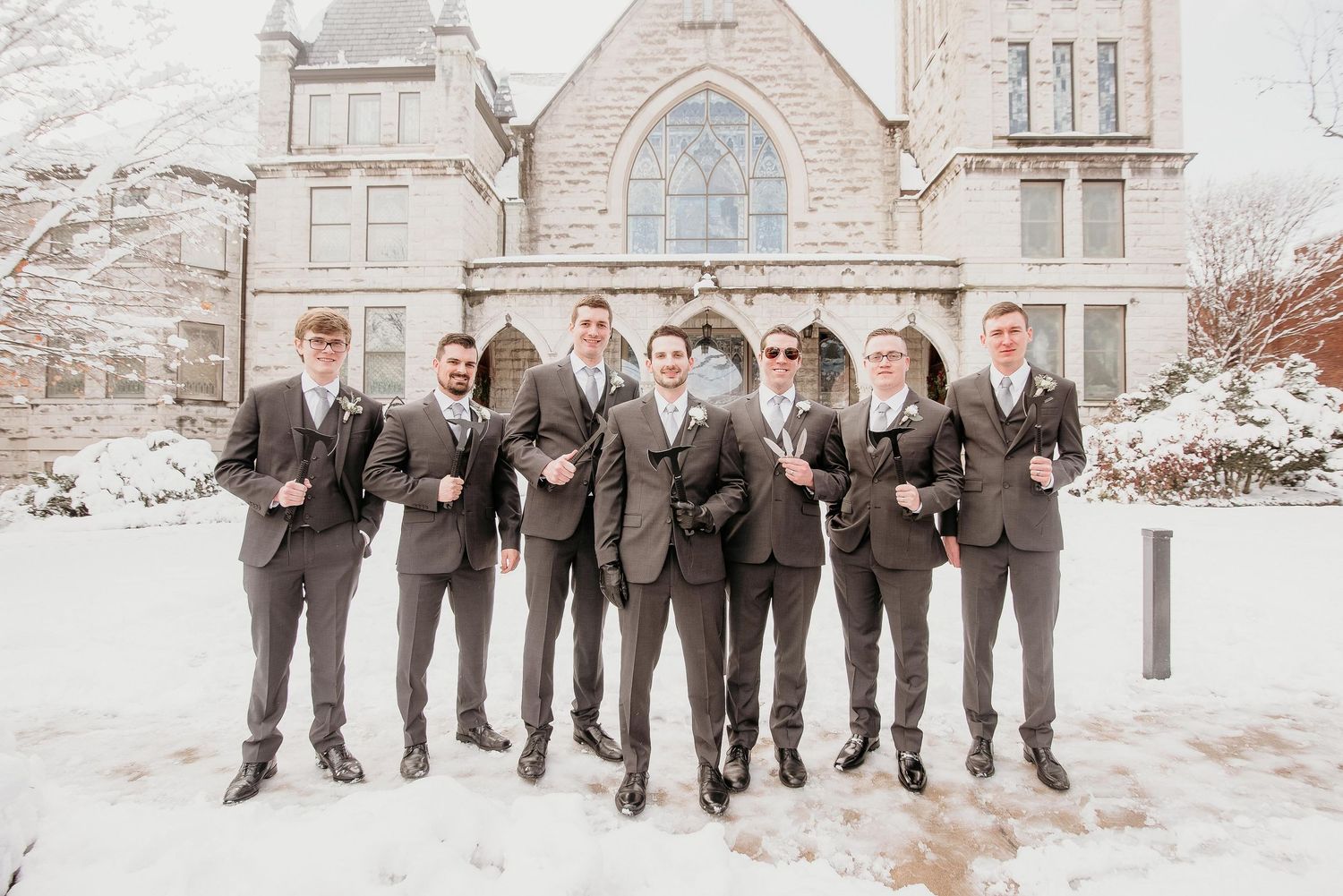 Groom and groomsmen in gray suits posing in snow outside a stone church on a winter wedding day.