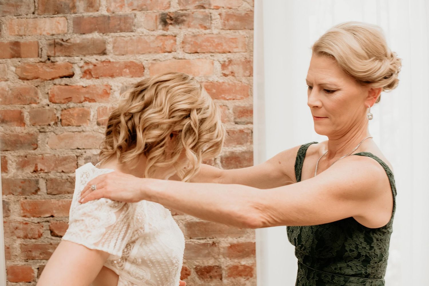 A woman in a green dress fastens the back of a bridal gown against a rustic brick wall backdrop.