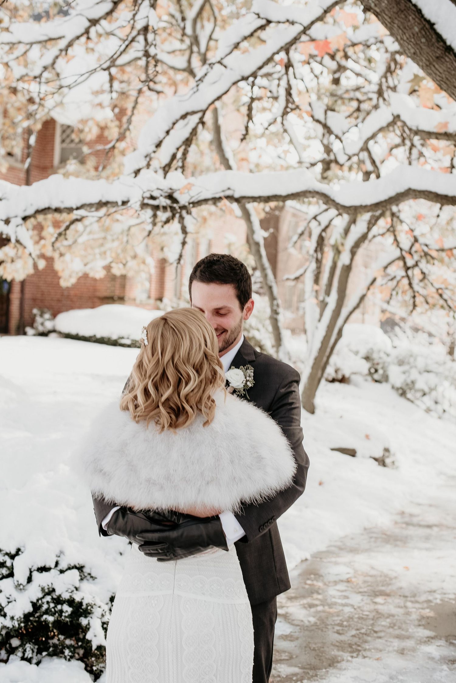Bride and groom embrace in snowy winter wonderland, bride wearing white fur stole and groom in gray suit.