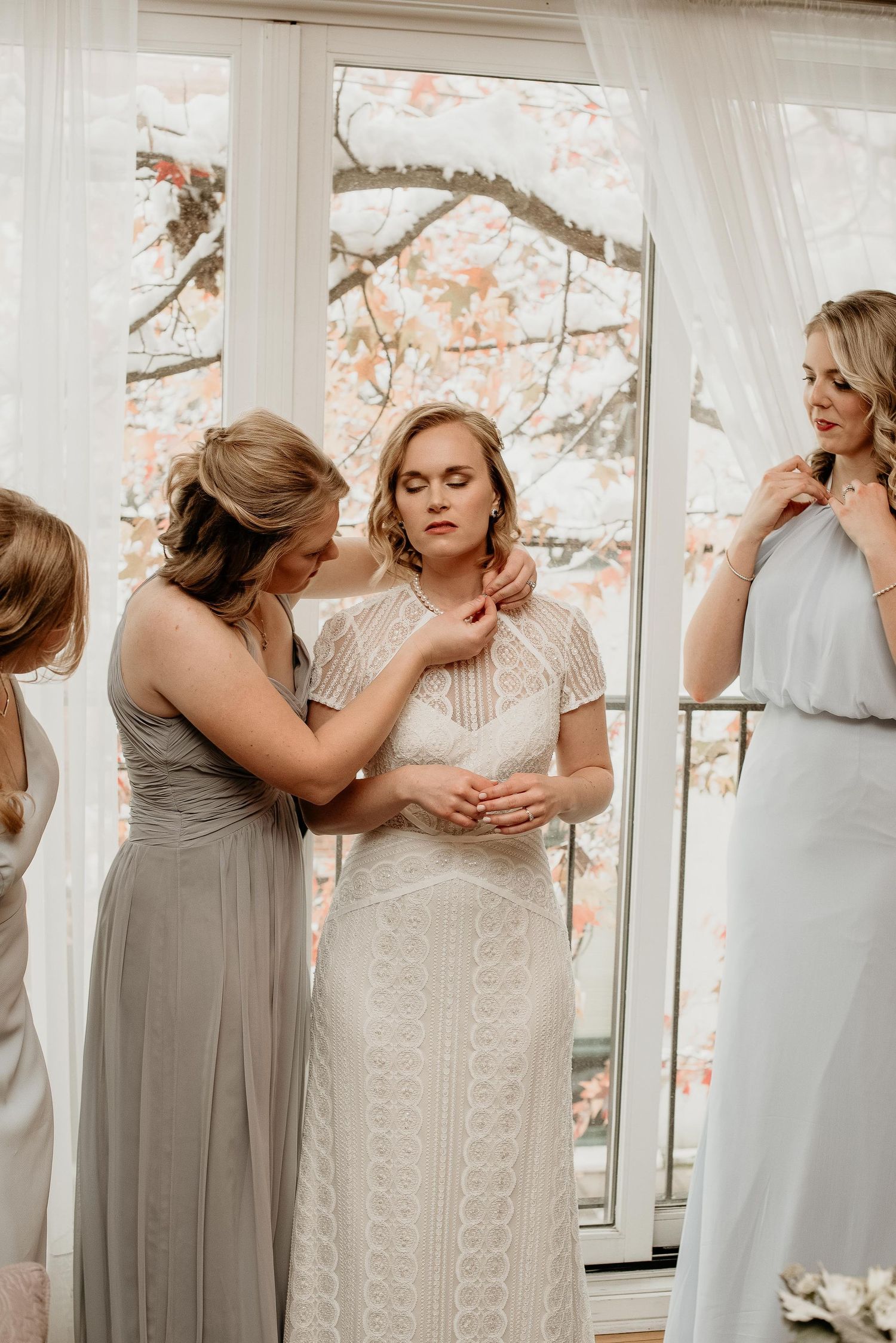 Bride in white lace gown gets jewelry help from bridesmaids in gray dresses near bright windows with floral backdrop.