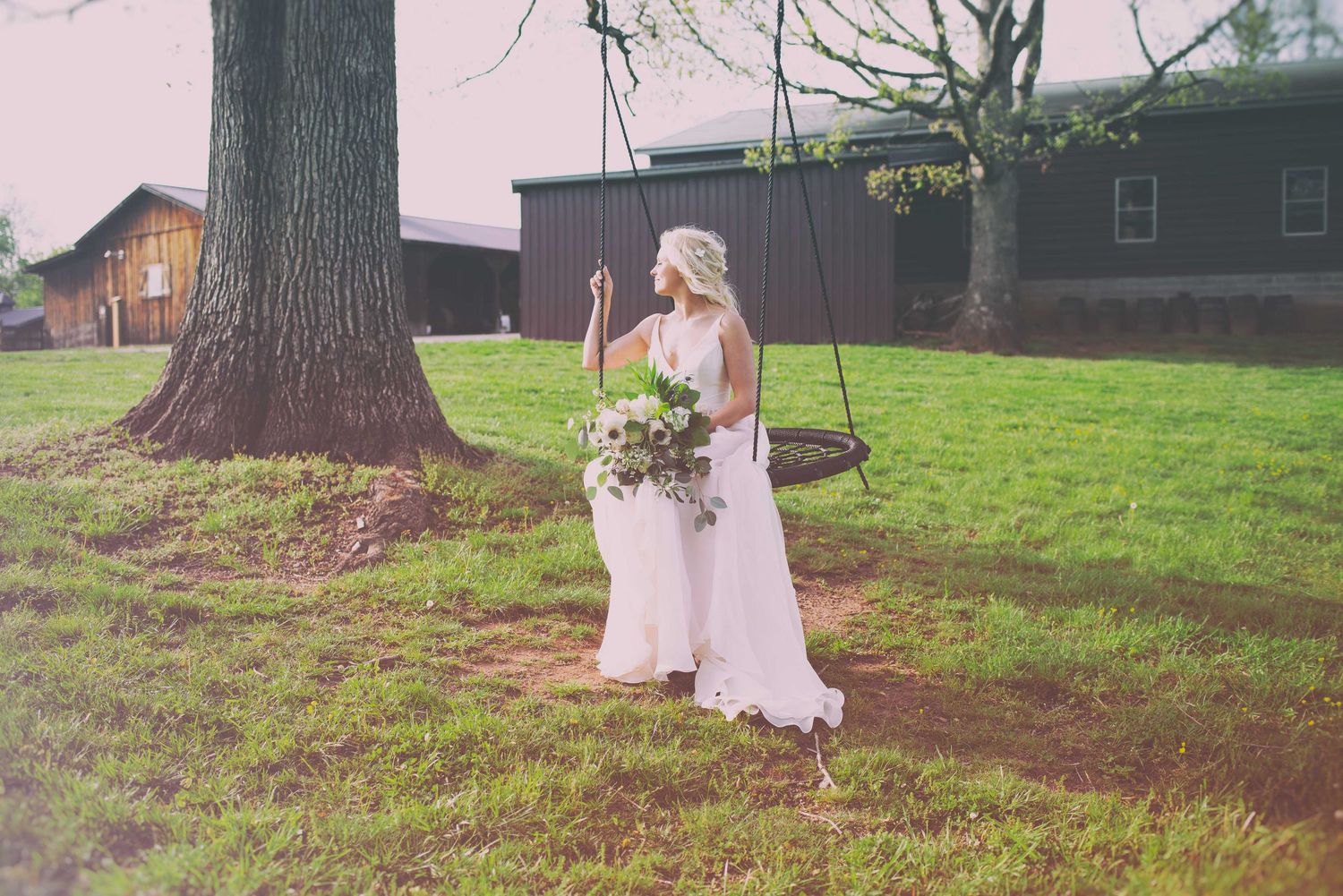 Bride in white gown holding bouquet stands by tree swing at rustic outdoor wedding venue on green lawn.