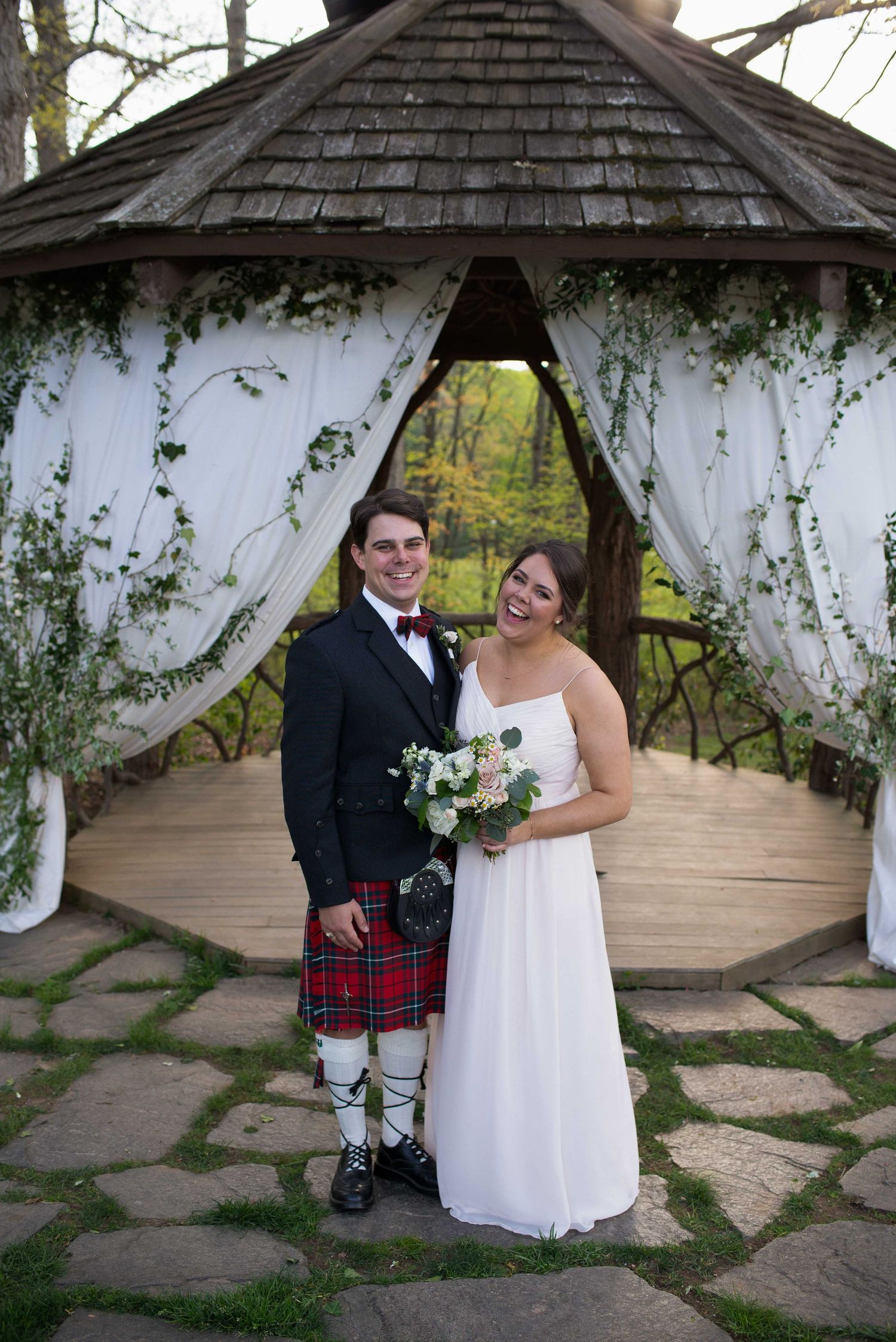 Smiling bride and groom in tartan kilt pose under ivy-draped gazebo with white curtains on stone path.