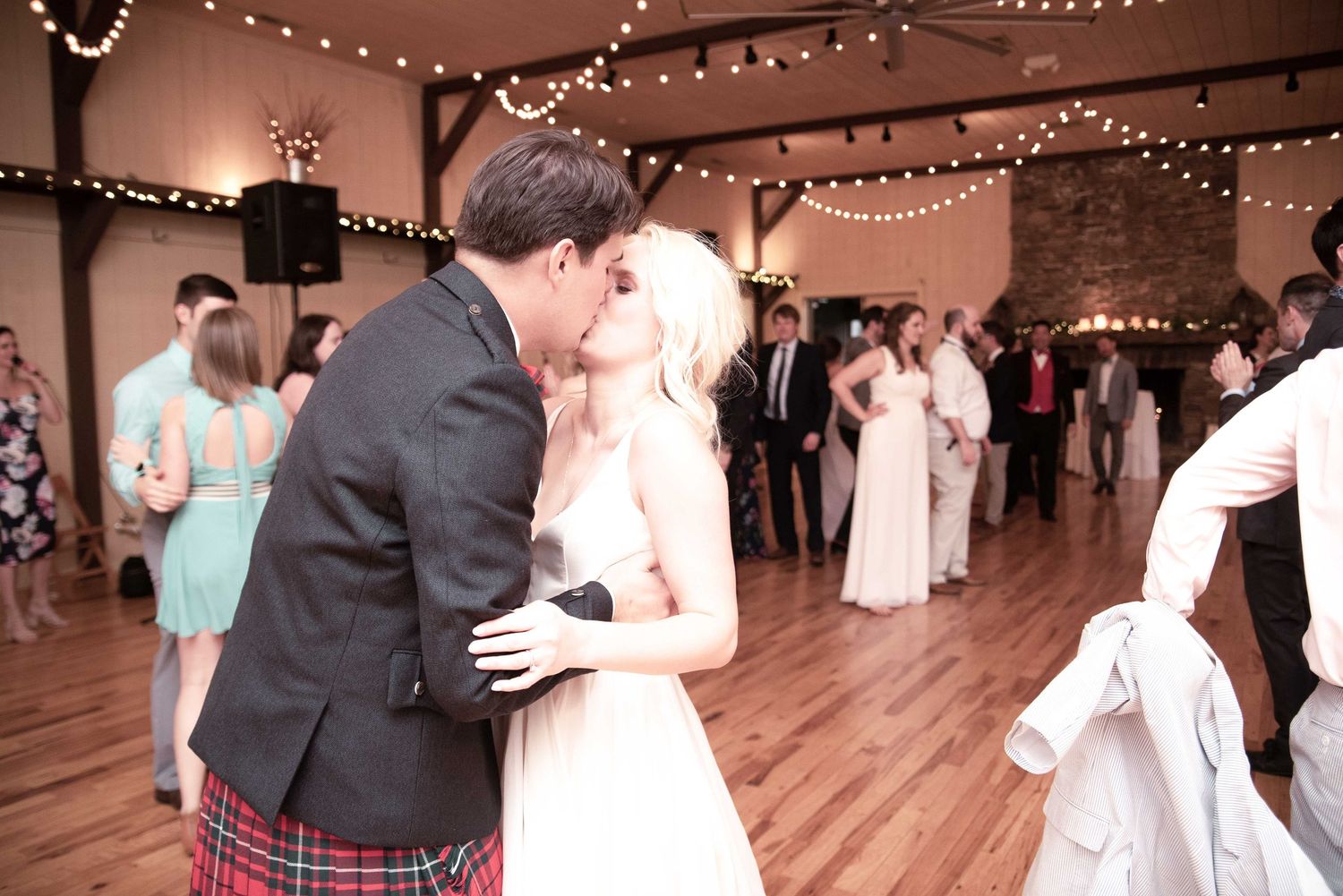 Bride and groom share a first dance kiss at wedding reception with string lights and guests in background.