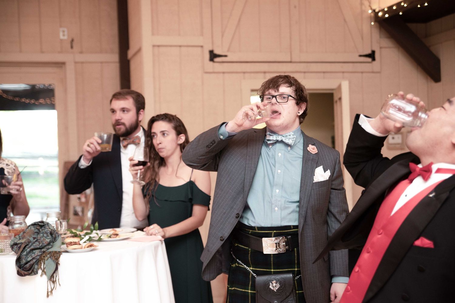 Wedding guests in formal attire toasting with drinks at a barn reception, man in kilt and bow tie.