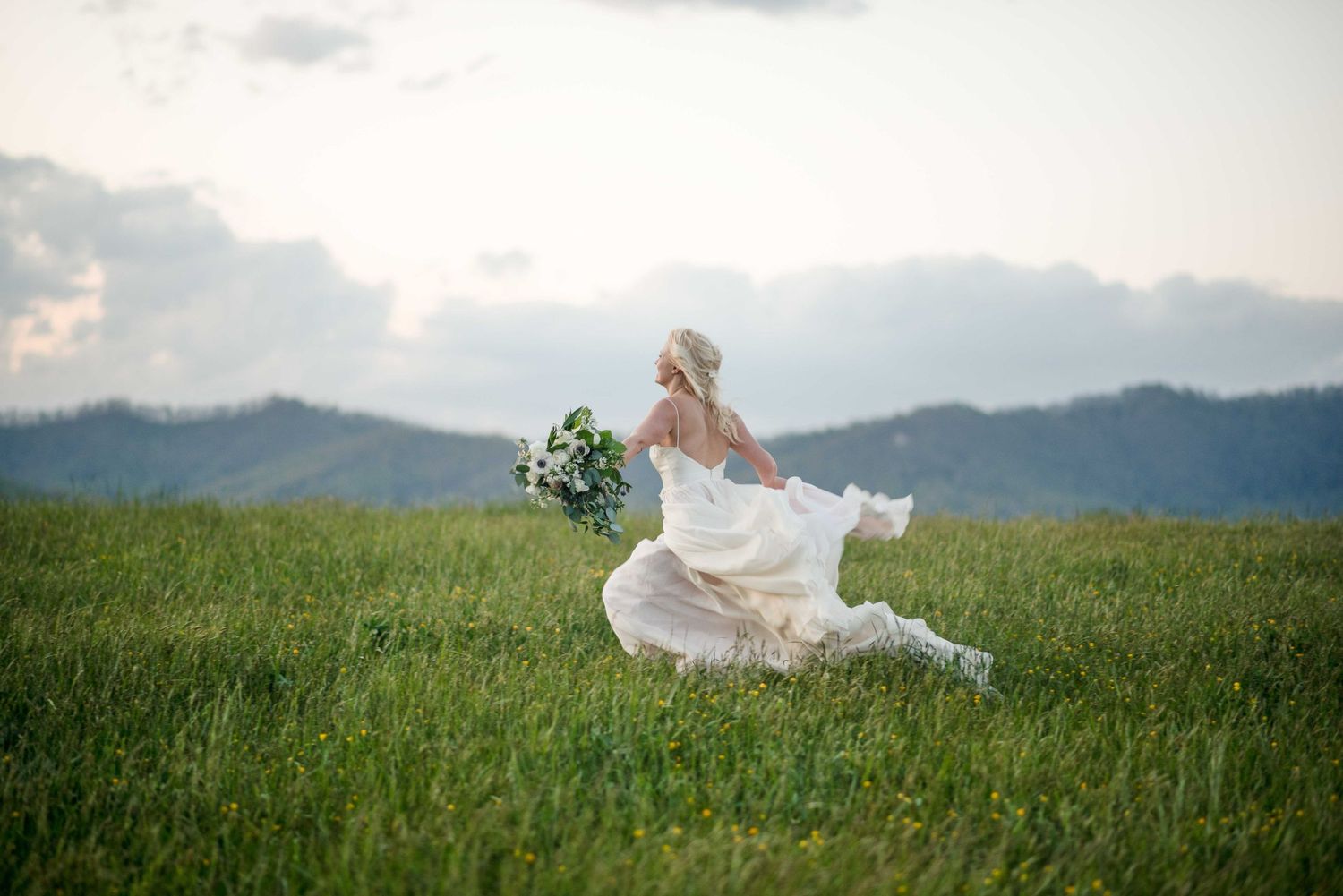 Bride in flowing white gown running through green meadow holding bouquet with mountain backdrop.