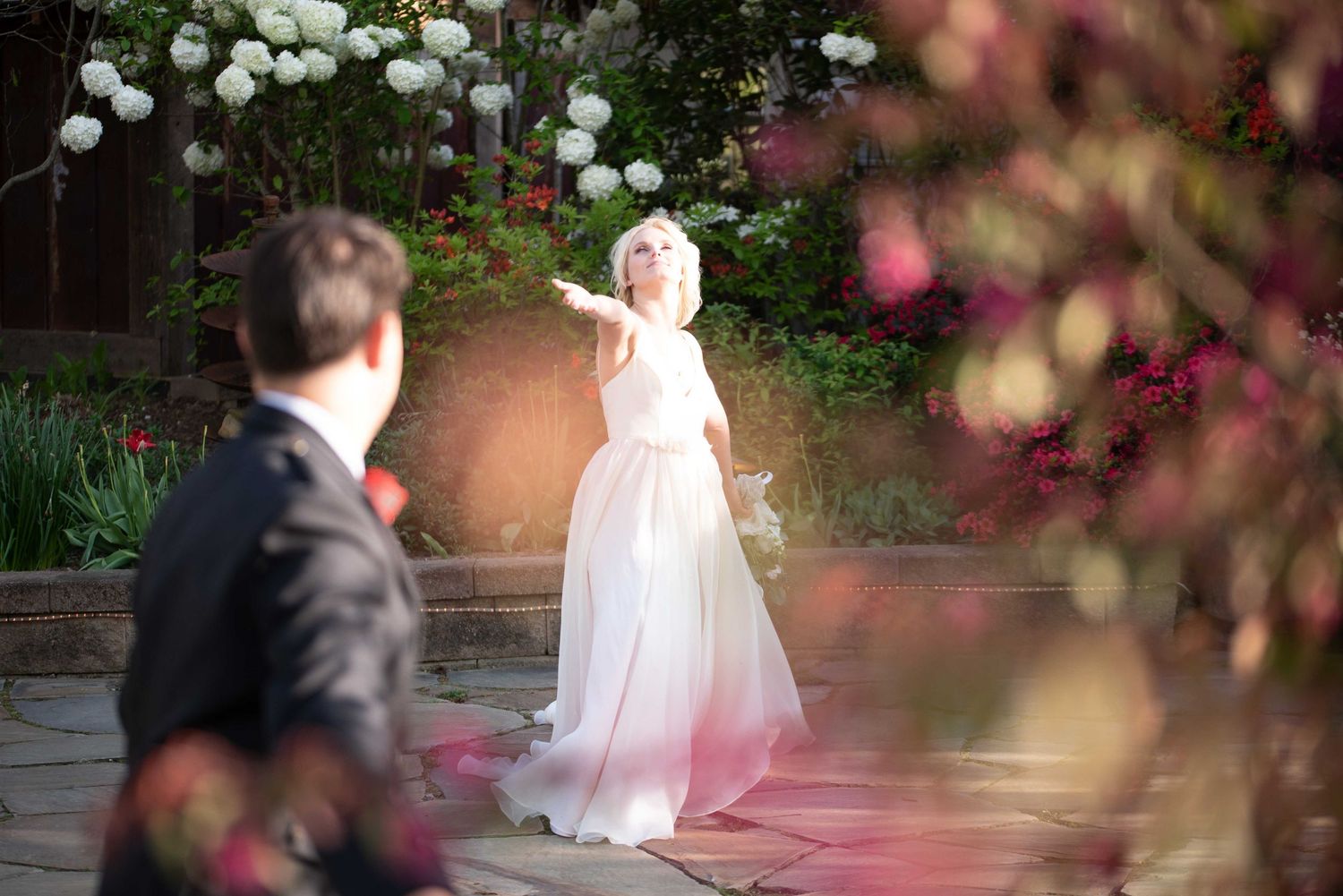 Bride in white ombre gown gestures outdoors amid blooming flowers and lush greenery at elegant garden wedding.