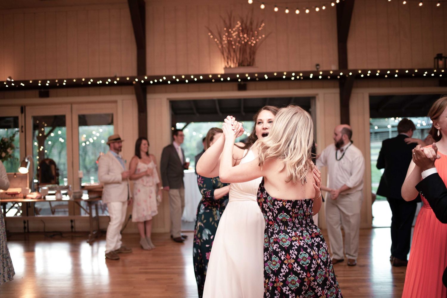 Two women dancing together at a rustic wedding reception venue with string lights and guests in background.