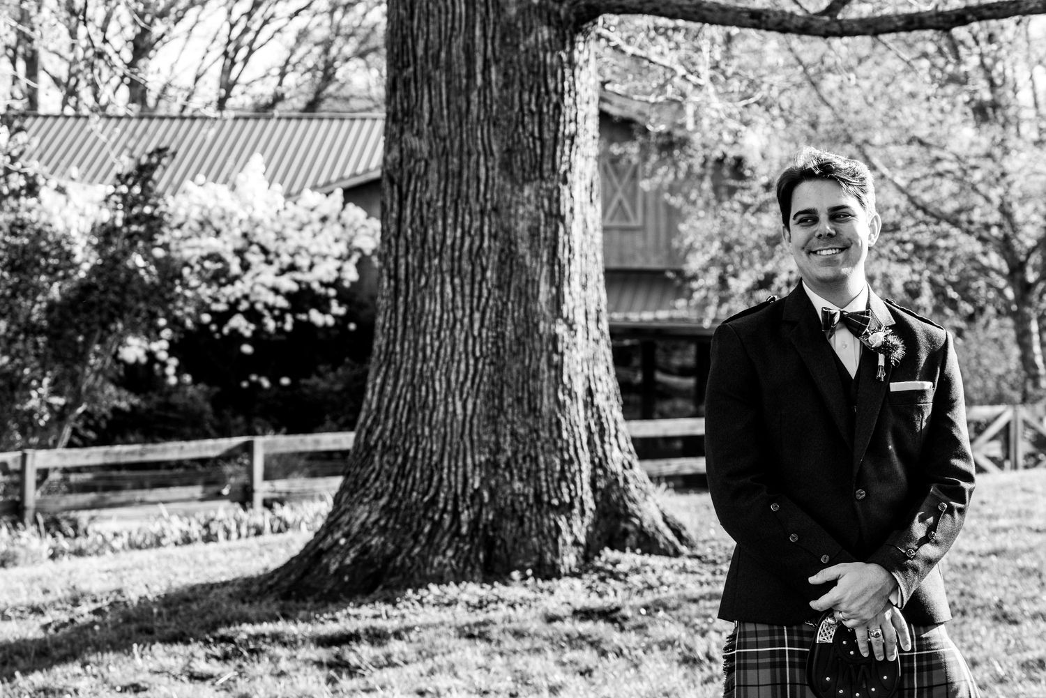 Man in traditional Scottish kilt and jacket poses by a large tree in a garden, black and white wedding photo.