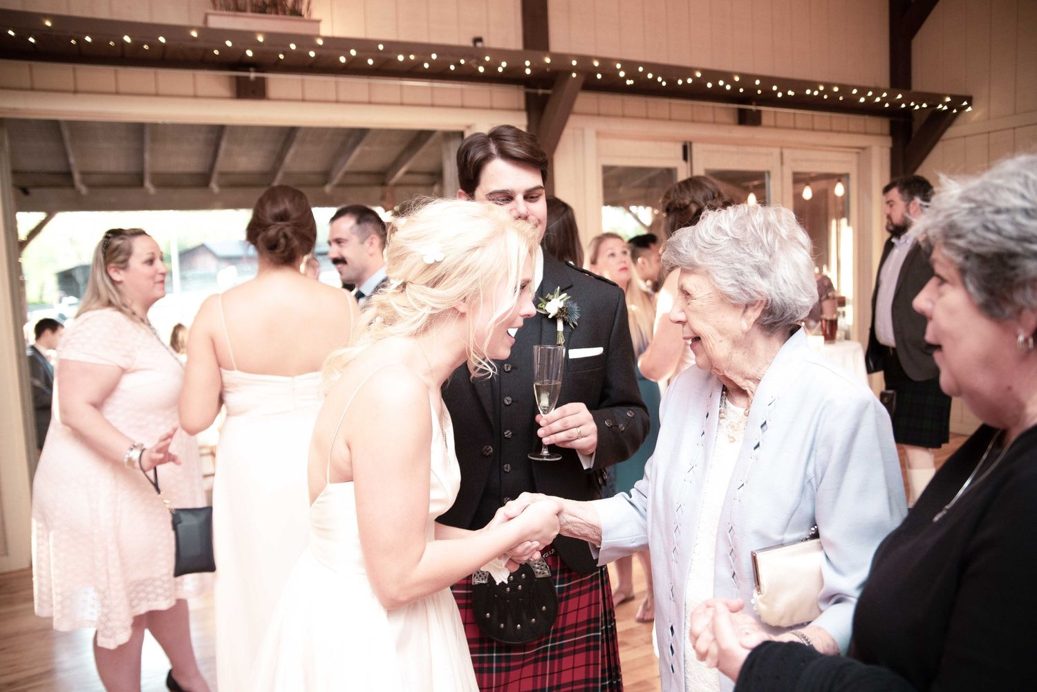 Bride and groom in Scottish kilt greeting elderly guest at elegant barn wedding reception.