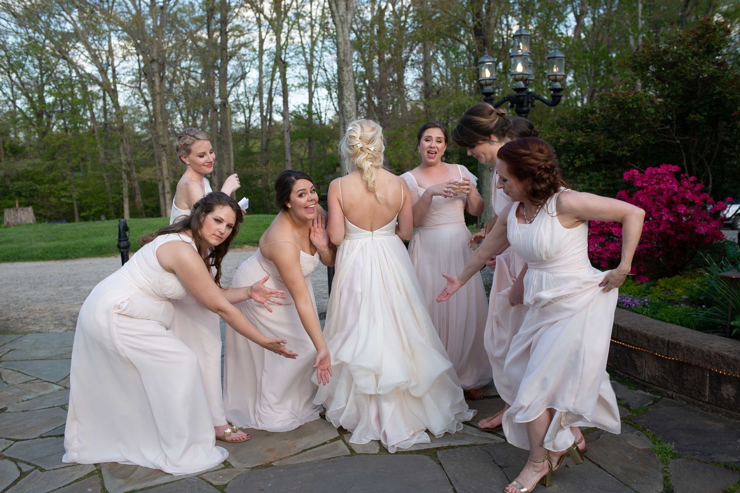 Bride surrounded by bridesmaids in blush gowns adjusting her white wedding dress outdoors.